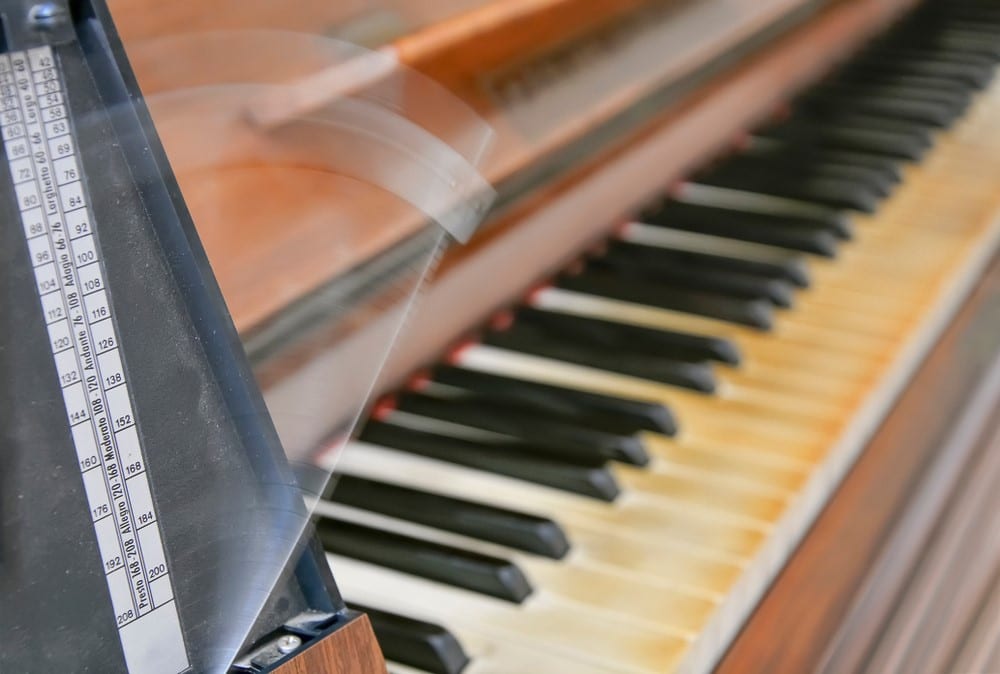 Old wooden retro metronome with piano on background. Musical equipment. Instrument to measure the music time speed or Tempo. Metronome swings its pendulum whom motion produces the classical tick sound